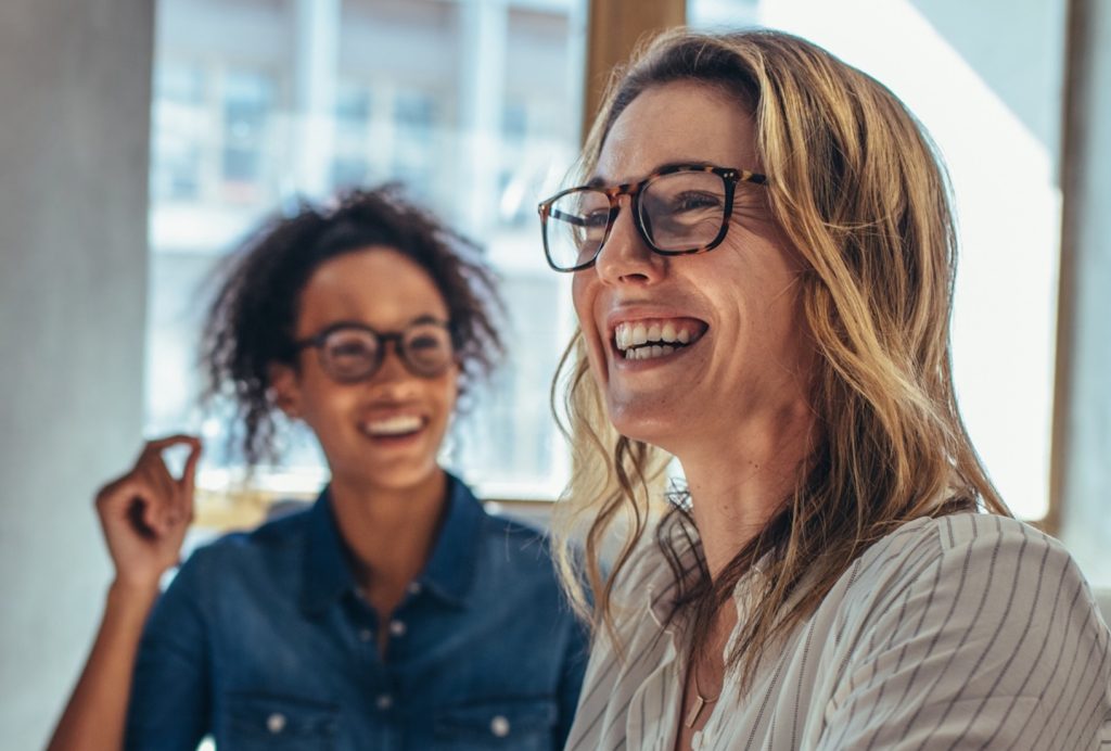 Two smiling businesswomen wearing glasses in a modern office environment, engaged in a positive team discussion, representing collaboration, leadership, and a supportive company culture.